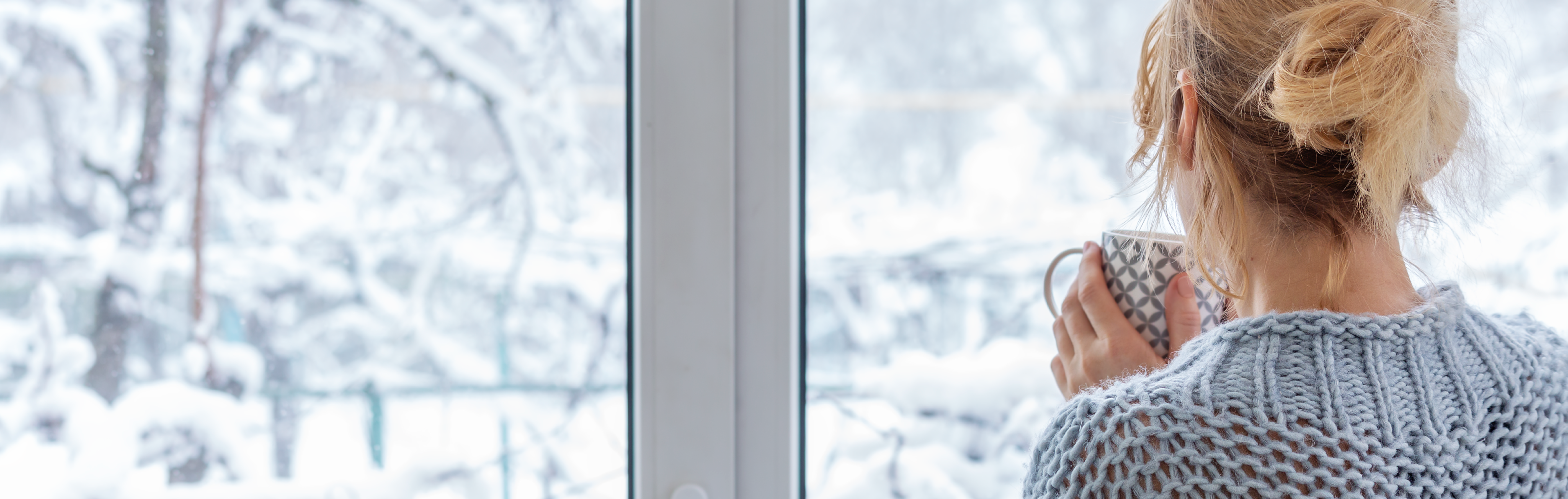 Woman holding coffee looking out window at snow